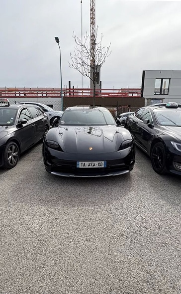 Porsche Taycan noire : voiture électrique entre taxi et chantier Luxueuse Porsche Taycan noire (plaque TA-AΓA-XΩ) garée entre deux voitures dont une Tesla taxi, devant un chantier sous ciel gris.