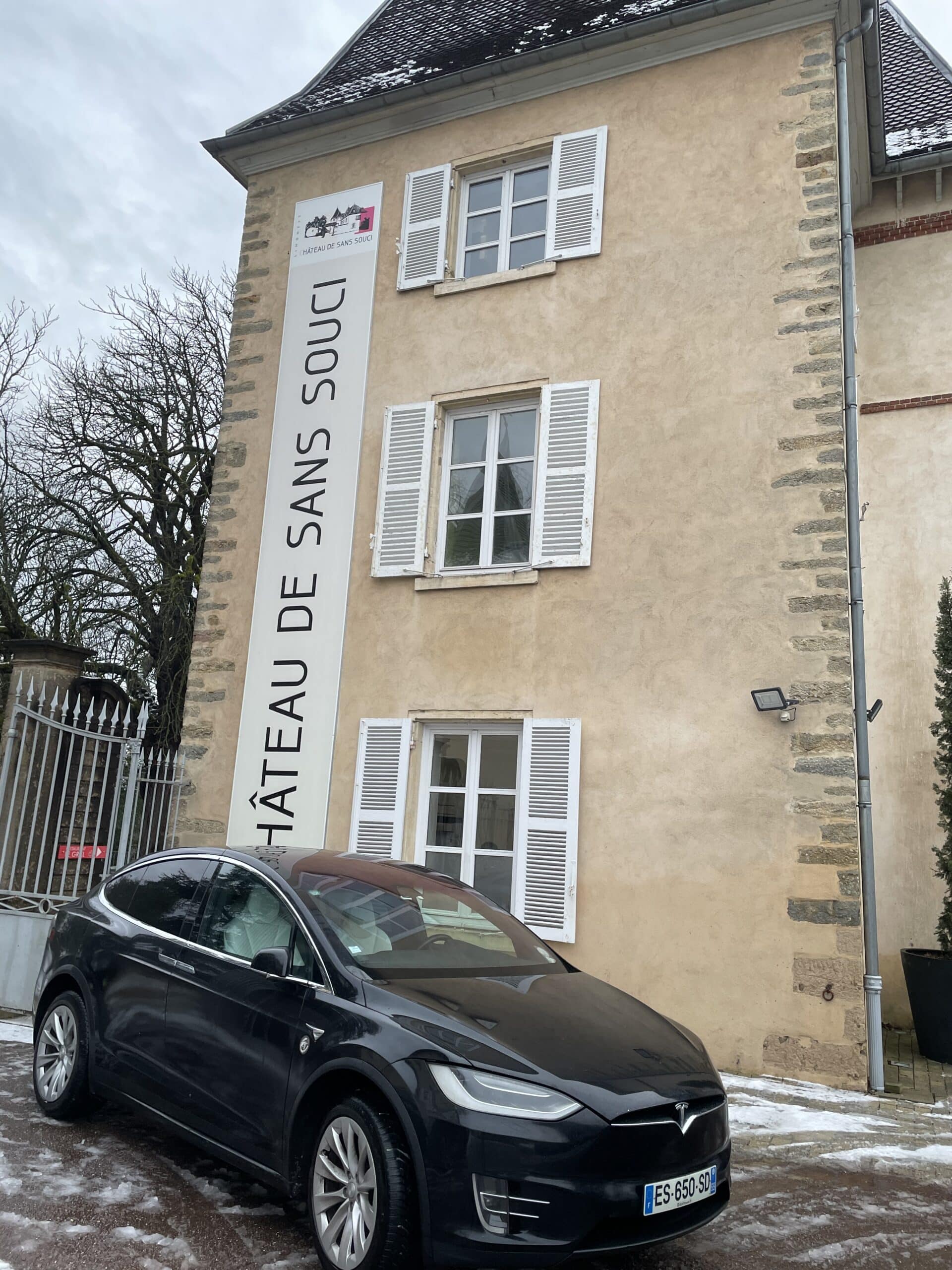 Tesla Model X noire garée devant le Château de Sans Souci Tesla noire garée devant le Château de Sans Souci, bâtiment historique avec volets blancs et bannière verticale.